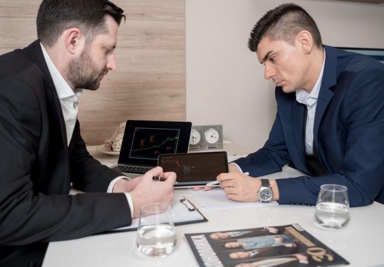 Two businessmen in suits discussing cryptocurrency trends using digital devices in an office setting.